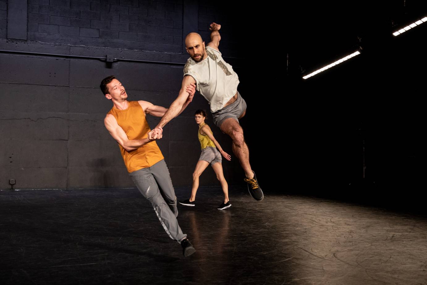 One man partners another in a stage leap as a woman looks on in the background